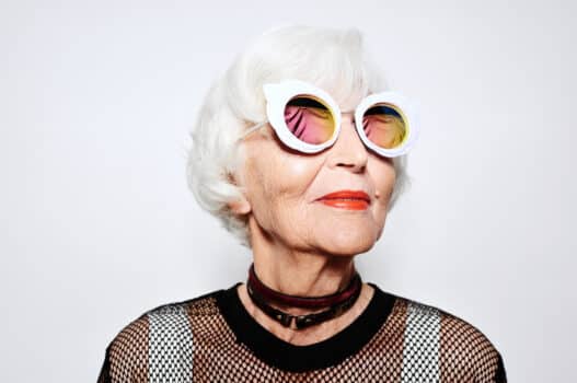 Close-up portrait of beautiful elderly woman in trendy clothes and sunglasses looking to the side and smiling against white wall background